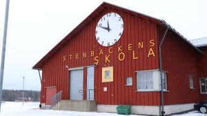 Stenbackens skola. En röd trähusfasad med texten Stenbackens skola uppristad på väggen med stora bokstäver. Ovanför texten en stor klocka.