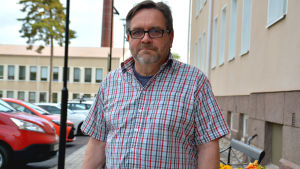 Samuel Nissfolk menar att resenärerna inte kommer att märka av någon stor förändring. Man i rutig skjorta står vid en gammal sjukhusbyggnad. Bilparkering i bakgrunden.