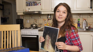 Sabina visar hur ärret såg ut till en början. Nu syns det inte alls under hennes tjocka hår. Sabina Granbacka sitter vid ett köksbord och håller i en bild som visar operationsärret som hon har på bakhuvudet.