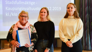 Fullmäktiges ordförande Tarja Rinne (sfp) tog emot en namninsamling som överräcktes av Anna-Maria Mårtensson och Wilma Karlgren. tre kvinnor, två yngre och en äldre står framför en digital skärm