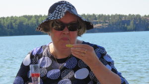 Ingen Invigning utan flädersaft och salta snittar. Nina Bergman njuter i skuggan. En lite äldre kvinna i solhatt och sommarklänning som dicker bubbel och äter salta snittar. Hav och sol i bakgrunden. Sommar.