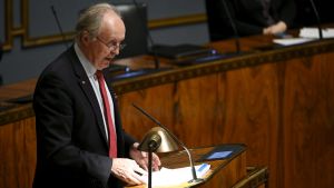 SDP:s Aki Lindén sa att det i praktiken är omöjligt att stifta en lag som gör det möjligt att bötfälla personer som inte använder munskydd. Aki Linden står i riksdagen och talar.