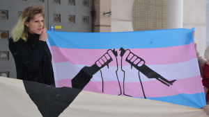 Elsa Tauria blir straffad för att hon vägrar göra militären och civiltjänstgöring. Elsa Tauria håller i en blå, rosa och vit banderoll med en bild av ett svart vapen som bryts i två delar.