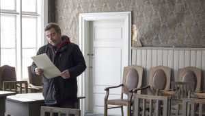 En del av rättegången hölls också på Tingshuset i Jakobstad. Här ser du Johan Strömberg, som spelat Pirilö-Karl i pjäsen "Rättegång på gång". Johan hörs också i podden. En man står i ett gammal hus. Det är ett stort rum med gamla buckliga tapeter. Längs med väggen står gamla stolar av trä. Han läser på ett papper.
