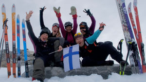 Ett glatt gäng som skidat över Grönland. Fem kvinnor iklädda tjocka vinterkläder. Sitter och hurrar och vifta med Finlands flagga. De befinner sig på Grönland, i ett snölandskap.