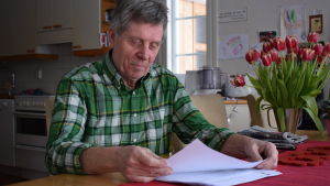 Cay Blomberg ser många brister i Balans-2020 kalkylerna. Cay Blomberg sitter med papper i handen vid sitt köksbord. Grönrutig skjorta på sig och en bukett tulpaner på bordet.