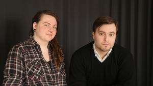 En ung kvinna med långt rödbrunt hår står bredvid en ung man som lutar sig mot ett bord.