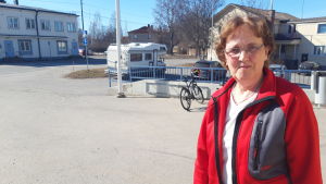 Helen Forsbacka En äldre dam med röd tröja står på en asfalterad gårdsplan. Hon har medellångt mörkt hår och glasögon.