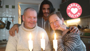 Frank Hellgren, Ahmed Aljuaifari och Pia Rousku poserar bakom en ljusstake med tre tända ljus i familjens kök