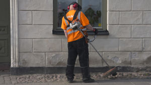 En person står med ryggen till och trimmar gräs utanför ett stenhus med en trimmer. Orange skjorta, reflex, hörselskydd. Sommar.