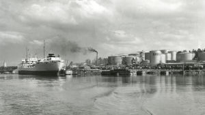 Oljehamn: Oljecisterner intill strand, stort lastfartyg vid brygga.