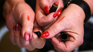 Läppstiftstestning. Tre händer visar upp rött nagellack på naglarna och streck av ett rött läppstift på tummen.