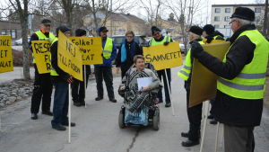 En kvinna i motoriserad rullstol kommer fram bland en liten grupp personer som håller gula plakat med sloganer mot en kommunsammanslagning.