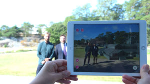 Företagarna Ilkka Rautio och Jacob Roos fotograferas via appen i badhusparken i Hangö. I verkligheten står de vid en grön gräsmatta, men via applikationen kommer en kopia av det tidigare badhuset med i bilden bredvid männen. Två män står bredvid varandra och fotograferas med hjälp av en mobilplatta. De befinner sig i en miljö med förstärkt verklighet.