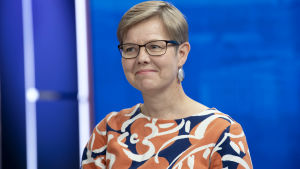 Miljöminister Krista Mikkonen (Gröna) i närbild i en studiomiljö. Hon är i närbild och ler snett mot kameran i en färggrann skjorta. Studioväggen är blå.