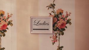 Kyltti, jossa lukee "Ladies" kukkatapetoidulla seinällä.