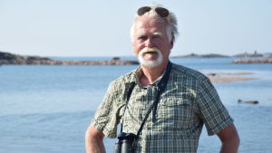 Mikael Kilpi har under många år forskat i ejdern. Nu är han pensionär men han studerar fortfarande fågeln. En man står med en kikare runt halsen. I bakgrunden syns hav och öar. Mannen är vithårig och har vitt skägg. På huvudet har han solglasögon