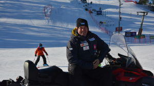 Jari Lehtinen är på bra humör i det soliga vintervädret. Jari Lehtinen, en man med svart jacka och svart mössa, sitter på en snöskoter. Bakom honom kommer en utförsåkare i röd jacka ner för skidbacken.