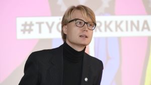 Debattören Pontus Purokuru talade för möjligheten att vägra jobba men ändå arbetslöshetsersättningar. Pontus Purokuru talar framför finsk skylt: Arbetsmarlknad