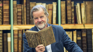 Gunnar Bjursell är intresserad av hur kultur och musik påverkar vår hjärna. En man iklädd blå kavaj står framför en bokhylla. Han har grått hår och skägg, och håller en bok i handen.