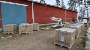 De 164 solpanelerna är fortfarande inpacketerade på lastpallar men snart är de redo att lyftas upp på taket. lastpallar med inpacketerade solpaneler intill ett stort rött ridhus. En skylift intill paketen.