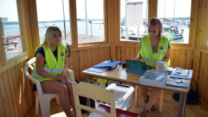 Wilma Rehnberg och Emilia Sundman hjälper båtfolket. två flickor sitter invid fönster i hamn.