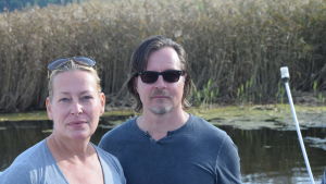 Eeva Moore och Juha Salminen. En kvinna och en man står på en båt vid en vassvik.