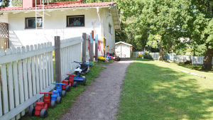 En bild på en gård där barntrampbilar står uppradade längsmed ett staket. På bilden syns hus och gräs