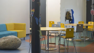 Skolans aula står till det provisoriska eftisets förfogande. Eftislokal i Nordsjös lågstadieskola. Färgglada stolar runt bord och en soffa längs väggen.