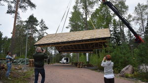 Det stora lyftet dokumenteras av Ingå kommuns informatör Kristoffer Nöjd, byggbolagets vd Paul Lynch och husägare Marjatta Lyytikäinen. Och jo, av Yle Västnylands reporter. Ett tak lyfts upp från marken med lyftkran. Några människor fotograferar.