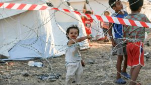 Sammanlagt omkring 600 personer har isolerats, säger Skaarup, eftersom även familjemedlemmar till smittade måste sitta i karantän. Barn som leker bakom taggtråd i flyktinglägret i Kara Tepe.