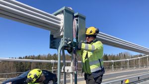 De grågröna stöden är det nya hängstaget. Under natten tas den tjocka grå vajern mittemellan dem bort. Två män iförda arbetskläder byter ut ett hängstag på Rävsundsbron en solig dag.