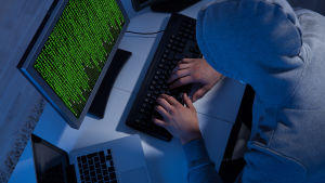 Person iklädd munkjacka sitter vid en laptop och en dator med en massa gröna tecken på skärmen.