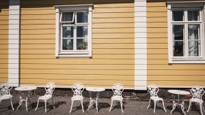 En solig sommardag kan du också njuta av ditt kaffe på utsidan av Handelshuset. På utsidan av handelshuset. Vita bord och stolar i solskenet framför handelshusets gula vägg.