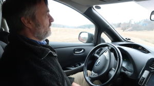 Ljudmiljön i bilen är tyst. Kurt Andersson sitter inne i sin elbil. Utanför ett landskap med åker i gråmulet vårväder.