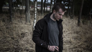 Foto: Niklas Meltio David står i en skog och tittar ner mot marken med ett litet leende på läpparna.