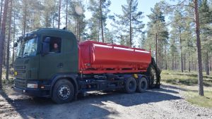 Den andra brandbilen rymmer 12 000 liter vatten. Den kan bara köra på vägar. En bild på en tankbil som fungerar som brandbil vid behov. Tankbilen är röd. Runt omkring den finns det tallskog. Tankbilen tillhör Försvarsmakten.