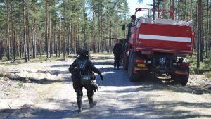 En del övningar kräver full utrustning trots hettan. En beväring går längs med en sandig väg i en skog. Beväringen har mycket utrustning på sig och går mot en militärisk brandbil som är röd. Man ser bakändan på bilen.