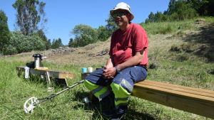 Anders Holmlund letar fynd på Slottsmalmen. En man i röd t-skjorta och arbetsbyxor sitter på en bänk. Han har en metalldetektor med sig och han sitter ute i naturen på ett område där man letar arkeologiska fynd.
