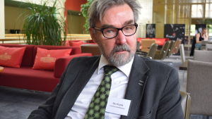 Naturresursinstitutets forskningsprofessor Ilpo Kojola. ilpo mKojola på Naturresursinstitutet stirrar stint in i kameran. Han har gråsprängt hår, grått skägg och svartbågade glasögon och är klädd i en grönprickig slips och grå kavaj.