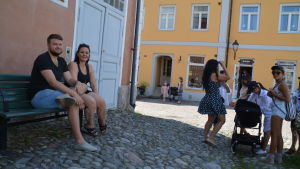 Maija och Olli-Pekka från Villmanstrand kämpade om en pausplats i skuggan. Två personer sitter på bänk intill husvägg och ler, en familj står nära bänken.