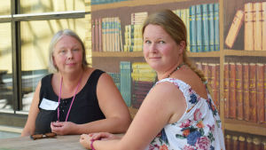 Mia Nylund och Henna Wilkman Två kvinnor sitter vid ett träbord. Den ena kvinnan har vänt sig om mot kameran så att man ser hennes rygg och den andra kvinnan sitter rakt i riktning mot kameran