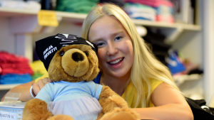 Sonja Laitamäki jobbar i foajén på sin dansskola. En blond ung kvinna ler och håller i en nalle som har en duk på huvudet där det står "Hurja Piruetti".