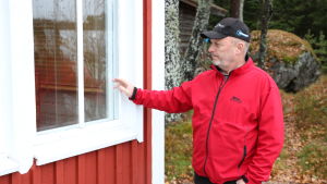 Här hade tjuvarna tagit sig in. En medelåders man med röd jacka och keps står intill en röd husvägg och pekar på ett fönster.