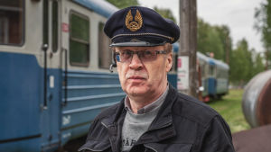 Ilkka Herpman. Vanhempi mies, jolla on päässään konduktöörinlakki. Taustalla sinisiä junavaunuja.