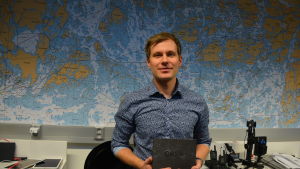 Conny Sjöqvist Ung man i blå skjorta håller upp en svart bok och står framför karta över skärgården