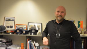 polisinspektör Måns Enqvist en ljus skäggig man i medelåldern klädd i svart skjorta. Står leende och lutar sig mot hylla med polishattar av olika slag