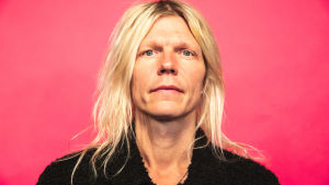 Jori Sjöroos katsoo kameraan lähikuvassa pinkin taustan edessä.