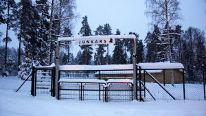 På Junkars har det ordnats danser för Svartåborna. En grind där det står Junkars. I bakgrunden syns en gul danspaviljong. Det ligger mycket snö på marken.