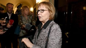 Vänsterförbundets Annika Lapintie, grundlagsutskottets ordförande, inför utskottets möte. En kvinna i spräcklig kavaj utanför en stor trädörr med en skara journalister kring sig.